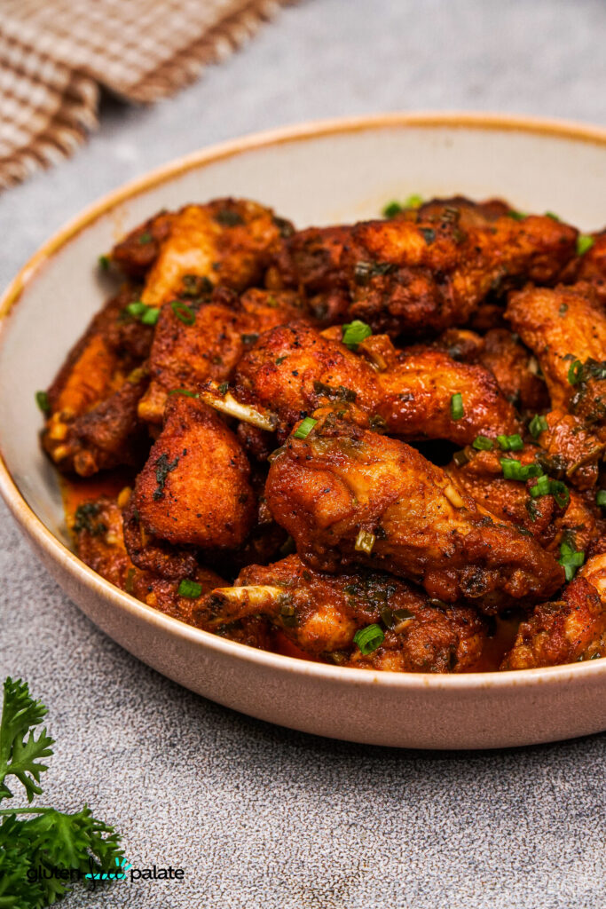 A close-up of crispy cowboy butter chicken wings in a ceramic bowl, coated in a smoky butter sauce and garnished with fresh herbs.