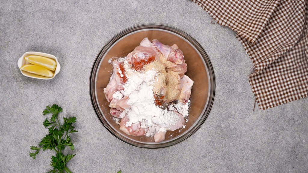 A glass bowl filled with raw chicken wings topped with cornstarch, paprika, garlic powder, salt, and pepper, ready to be mixed for seasoning.