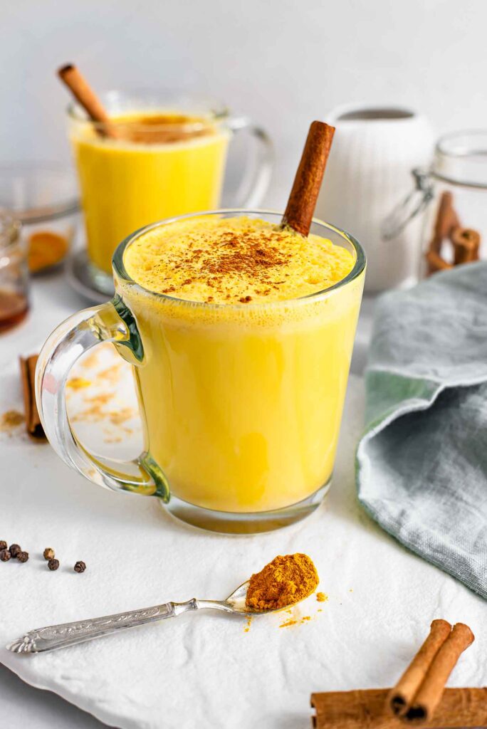 Turmeric Latte in a clear glass with spices surrounding it. Photo by tastythriftytimely.com.