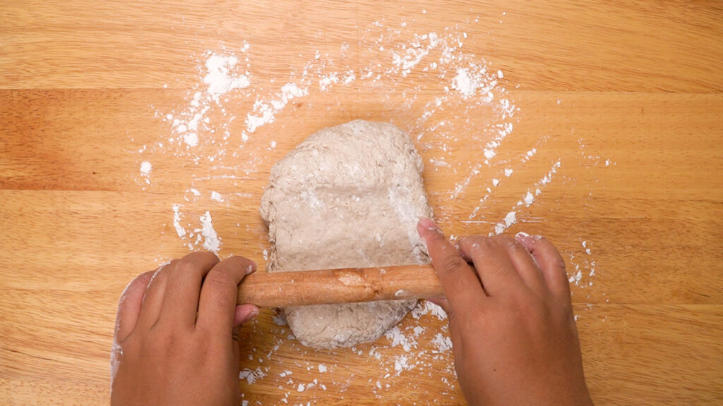 Hands rolling out sticky gluten-free ciabatta dough on a floured wooden surface.