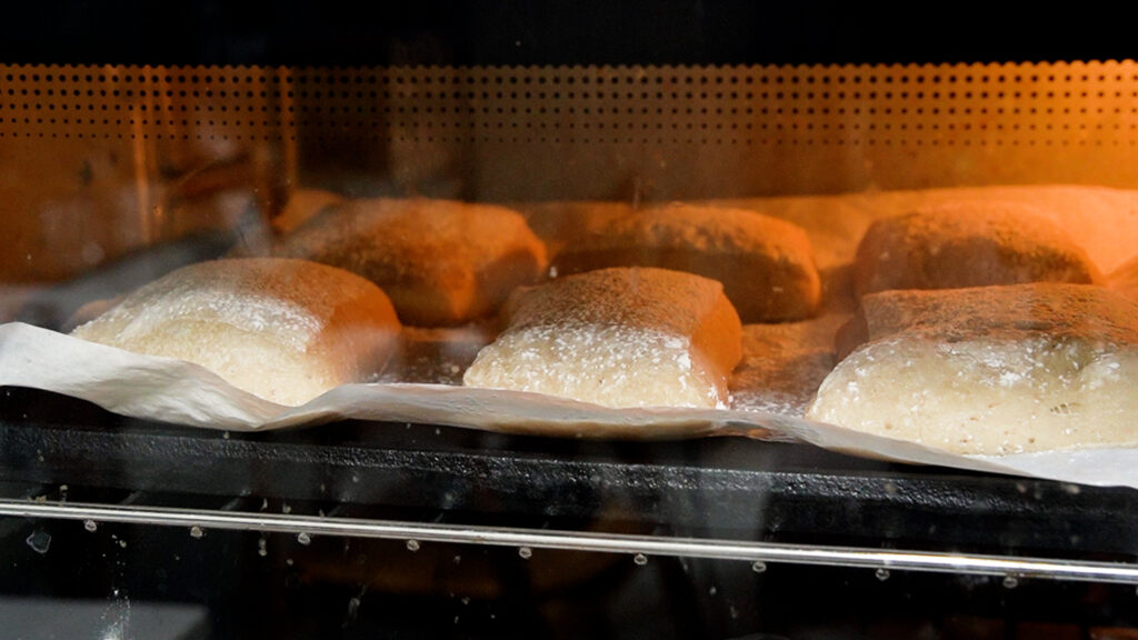 Gluten-free ciabatta rolls baking in the oven on a parchment-lined tray with steam.