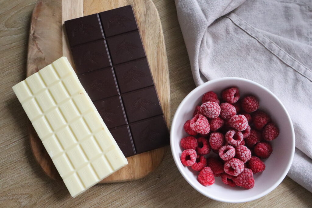 White chocolate, dark chocolate, and a bowl of frozen raspberries arranged on a wooden board.