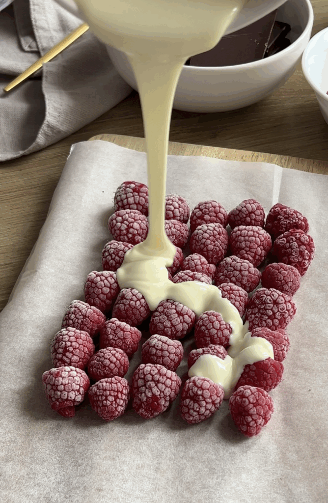 Melted white chocolate being poured over a layer of frozen raspberries on parchment paper.