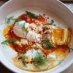 A bowl of Turkish eggs with poached eggs on creamy yogurt, topped with red chili butter and fresh dill.