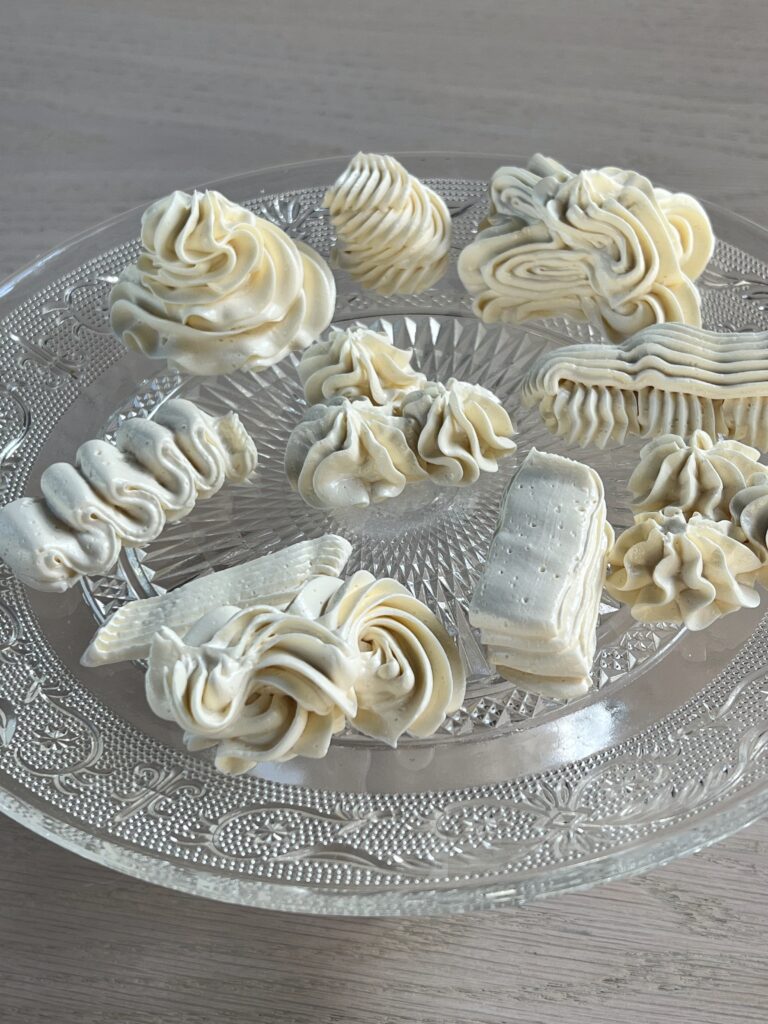 Italian meringue buttercream piped in different decorative styles on a clear glass serving plate.