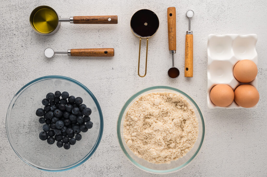 Almond flour muffins ingredients laid out on a countertop including almond flour, eggs, oil, honey, and blueberries