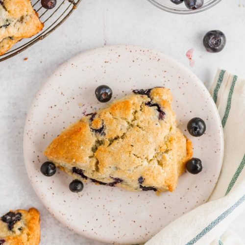 a blueberry scone on a white textured plate with blueberries and a towel